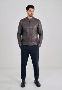 Mężczyzna stojący z rękami w kieszeniach, ubrany w ciemnobrązową skórzaną kurtkę, czarne spodnie i czarne buty na jednolitym jasnym tle.