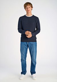 Marineblauer Kabelstrickpullover mit Rundhalsausschnitt, kombiniert mit blauen Jeans und weißen Sneakers. Weiche Textur mit gerippten Akzenten an den Bündchen und am Saum.