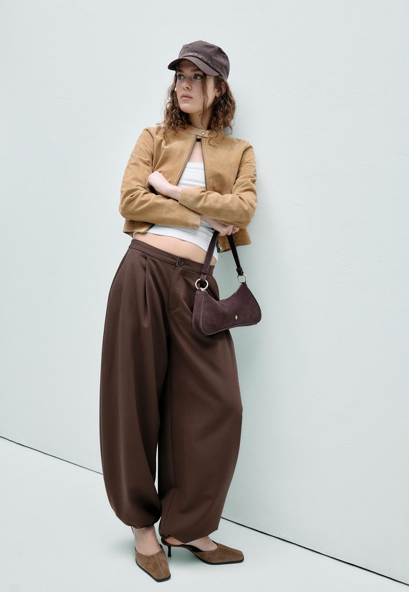 Jeune femme appuyée contre un mur blanc, portant un pantalon large marron, une veste courte couleur cuir clair, un haut blanc, une casquette noire, des talons marron et tenant un sac à main.