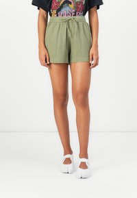Grüne Shorts mit einem elastischen Bund und Kordelzug, die einen lockeren Sitz und eine leichte Textur bieten, kombiniert mit weißen Schuhen.