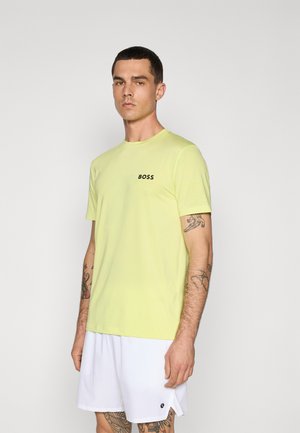 Lys gul kortærmet T-shirt med rund hals og sort "BOSS" logo på brystet, parret med hvide shorts, som viser tatoverede arme.