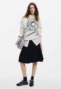 Suéter blanco con líneas abstractas negras, superpuesto sobre una camisa blanca con cuello. Falda negra, zapatillas negras y grises, sosteniendo un bolso plateado.