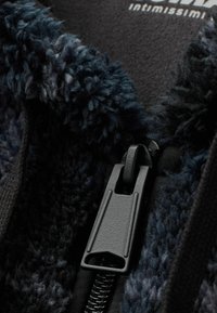 Modrá chlupatá tkanina s jemnou texturou, obsahující černý zip a kovové detaily. Materiál vypadá pohodlně a je vhodný pro vrchní oblečení.