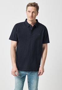 Marinblå pikétröja med korta ärmar, krage, treknappsknäppning och en subtil logotyp på bröstet. Bärs med ljusblå jeans.