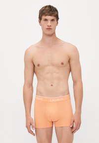 Jonge man die tegen een witte achtergrond staat, gekleed in perzikkleurige Lacoste boxershort, recht vooruit kijkend met een neutrale uitdrukking.