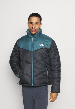 Giacca imbottita con una sezione superiore color turchese e una sezione inferiore nera, con un colletto alto e tasche con zip. Realizzata in tessuto leggero.