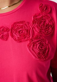 Chemise rose vif avec des roses en tissu texturé sur la poitrine et une chaîne en or visible au niveau du décolleté.