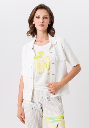 Vrouw draagt een wit kortermig jasje over een wit shirt met een gele citroenenprint, gecombineerd met lichtgekleurde broek met abstracte patronen.