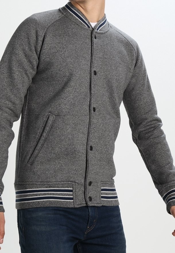 Homme portant une veste grise boutonnée avec des poignets et un col rayés, associée à un jean bleu, debout devant un fond uni.