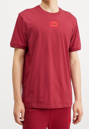 Mand iført en rød kortærmet T-shirt og matchende røde bukser, begge med små røde rektangulære "HUGO" logoer på brystet og låret.
