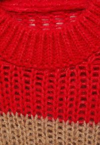 Top roșu din tricot, cu un guler texturat și căptușit, având un design cu model deschis. Mai jos, o secțiune solidă de culoare bej completează aspectul stratificat.