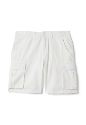Giovane uomo che indossa una polo blu navy, pantaloncini cargo bianchi, sneakers beige e un maglione azzurro chiaro legato in vita, in piedi davanti a uno sfondo semplice.