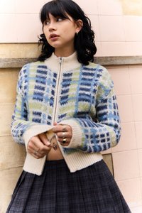 Cardigan tricotat cu un model în dungi în nuanțe de albastru și verde, având un guler crem cu fermoar și manșete elasticizate. Asortat cu o fustă cu carouri.