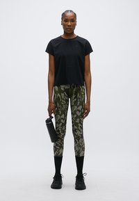 Μαύρο κομμένο αθλητικό μπλουζάκι, κολάν με πράσινη παραλλαγή, και μαύρα αθλητικά παπούτσια. Ένα μπουκάλι νερού κρατιέται στο αριστερό χέρι.