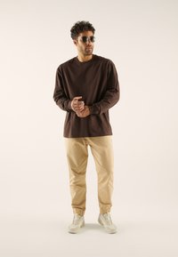 Chemise marron à manches longues, pantalon beige fuselé et baskets légères. Présente des lunettes de soleil rondes. Textures lisses et coupe décontractée.