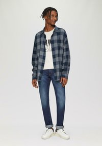 Marineblauw en grijs geruite flanellen overhemd over een crèmekleurig T-shirt met grafische print, gecombineerd met donkerblauwe jeans en witte sneakers. Casual look.