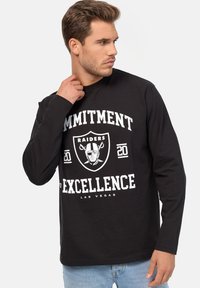 Schwarz langärmliges T-Shirt mit einem auffälligen weißen Grafikdesign, das "VERPFLICHTUNG ZUR EXZELLENZ" und ein Wappenlogo mit dem Gesicht eines Footballspielers zeigt.