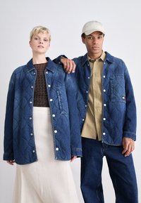 To personer iført denim-arbeidsjakker med kviltede mønstre, som har knappelukking og en lapp. En lysfarget skjorte er også synlig.