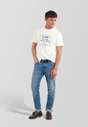 Joven con camiseta blanca con gráfico, vaqueros azules remangados, cinturón negro y zapatos marrón oscuro de pie frente a un fondo liso.