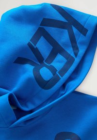 Blauwe hoodie van zachte stof, met grote, donkerblauwe grafische afbeeldingen op de capuchon en de borst. Gladde textuur met een relaxte fit.