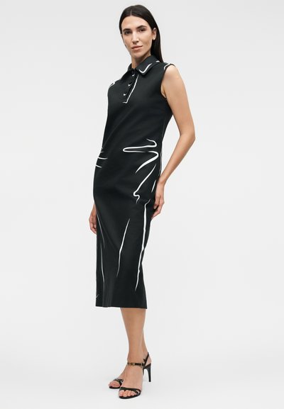 Γυναίκα που φοράει μαύρο midi φόρεμα χωρίς μανίκια με λευκές αφηρημένες γραμμές, γιακά, κουμπιά και μαύρα ψηλοτάκουνα σανδάλια, στέκεται πλευρικά.