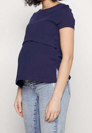 Mujer vistiendo una camiseta de maternidad azul marino de manga corta y jeans de mezclilla azul claro, de pie contra un fondo blanco liso.