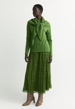 Frau trägt grünen Pullover mit passendem gebundenem Schal, grünen Spitzenrock und beige Absatz-Stiefeletten, steht vor weißem Hintergrund.