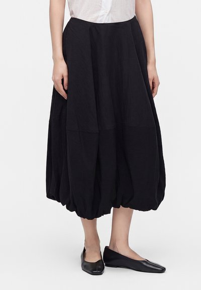 Femme portant une jupe midi noire à ourlet boule, une chemise blanche et des chaussures plates noires, debout devant un fond blanc uni.