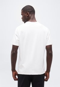 Άνδρας με την πλάτη, φοράει απλό λευκό κοντομάνικο μπλουζάκι και μαύρο παντελόνι, μπροστά σε απλό ανοιχτό φόντο.