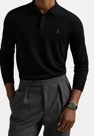 Mann trägt eng anliegendes schwarzes langärmeliges Polo und hoch taillierte graue plissierte Hose, ergänzt durch eine schwarze Armbanduhr mit Band am linken Handgelenk.
