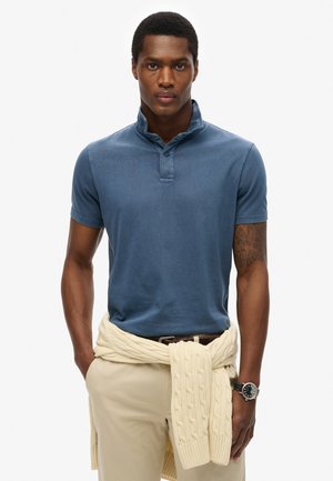 Polo shirt i blå bomuld, korte ærmer og en knapkrave. Cremefarvet striktrøje bundet omkring taljen, båret med beige bukser.
