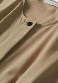 Chemise beige avec encolure ronde et une seule fermeture par bouton noir en haut, composée d'un tissu lisse avec des coutures visibles.