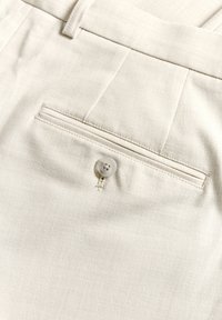 Beige byxor i tyg med en slät textur, med en bakficka som säkras av en ljusgrå knapp, kompletterat med subtila sömnadsdetaljer.