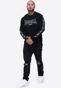 Czarna bluza z kapturem z logo "LONSDALE LONDON", ze ściągaczami na rękawach i detalami w designie na rękawach, w połączeniu z podartymi czarnymi dżinsami i białymi trampkami.