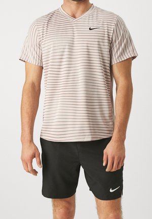 Man die een lichtbeige gestreept T-shirt met V-hals en zwarte Nike-short draagt, staat tegen een neutrale achtergrond.
