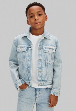 Jongen staat met handen in de zakken, draagt een lichtblauwe spijkerjasje en spijkerbroek over een wit T-shirt tegen een grijze achtergrond.