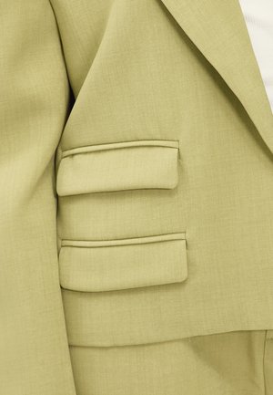 Blazer vert clair en tissu texturé, avec des revers crantés et des détails d'épaule rembourrés. Comprend trois accents rectangulaires sur les manches.