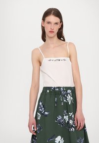 Top alb cu bretele subțiri, decorat cu broderie florala deasupra bustului, asortat cu o fustă verde ce are un model floral albastru și talie elastică.