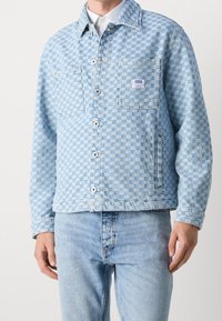 Light blue checkered denim jacket featuring a button closure, two front pockets, and a branded patch on the left pocket.
