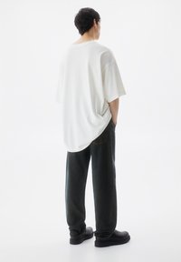 Maglietta bianca oversize con maniche corte indossata con pantaloni di denim scuro. Scarpe nere visibili. Sfondo minimalista e chiaro.