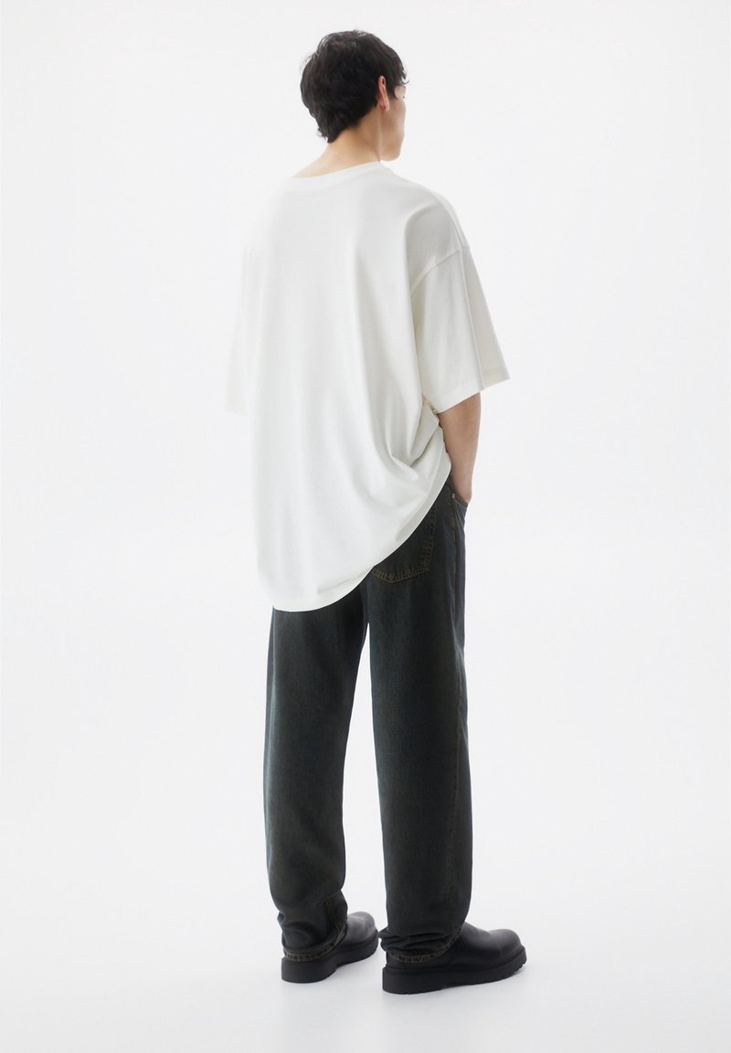 Maglietta bianca oversize con maniche corte indossata con pantaloni di denim scuro. Scarpe nere visibili. Sfondo minimalista e chiaro.