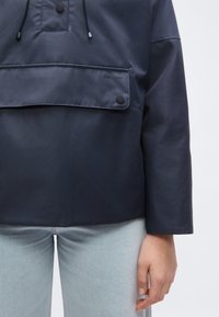 Μπλε ναυτικό μπουφάν Barbour με τσέπη με καπάκι και κουμπιά μπροστά, φορεμένο με ανοιχτό μπλε τζιν και ορατό χέρι στο πλάι.