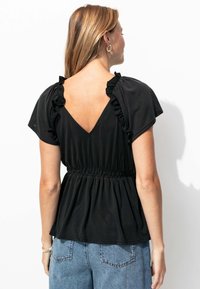 Le haut noir présente un dos en V, des manches courtes avec des accents en volants, et une taille élastique pour un effet froncé. Associé à un jean taille haute.