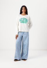 Felpa grigia chiara a maniche lunghe con testo verde "GAP SPORT 1969", abbinata a jeans larghi azzurri, indossata con scarpe nere senza lacci.