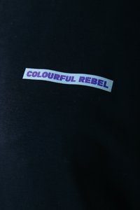 Tessuto nero con un'etichetta orizzontale riportante "COLOURFUL REBEL" in caratteri viola. La texture è liscia, con un motivo a coste visibile.