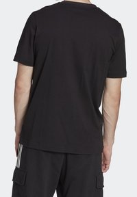 Zadní pohled na osobu v čistém černém tričku s krátkým rukávem a černých cargo kalhotách s bílými pruhy na bočních kapsách.