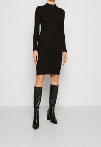 Abito lungo a maniche lunghe nero a coste con scollatura alta, silhouette aderente, abbinato a stivali neri in pelle con tacco e punta affusolata, fino al ginocchio.