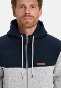 Chaqueta con capucha y cremallera que presenta una sección superior azul oscuro y una sección inferior gris claro. Fabricada con tejido texturizado y cordones de contraste.