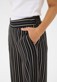 Černé a béžové pruhované kalhoty s širokým elastickým pasem a bočními kapsami. Materiál vypadá měkký, má hladkou strukturu a elegantně se splývá.