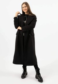 Zwarte wollen lange jas met grote revers, dubbelknopensluiting en voorzakken, gedragen over een grijs trui en gecombineerd met zwarte stevige laarzen.